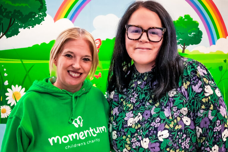 A Family Support Worker stands in front of a colourful mural with a rainbow and countryside scene on a children's hospital ward, together with a hospital staff member.