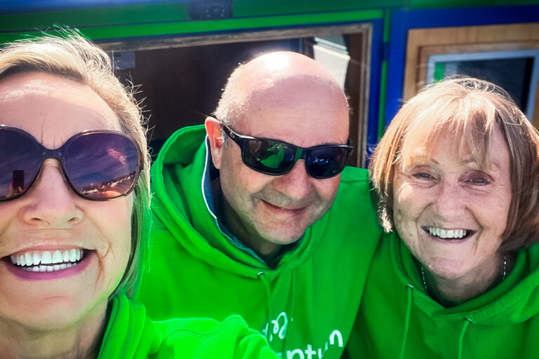 Three Momentum river boat volunteers, two wearing sunglasses, pose do a smiley selfie in front of the boat on a sunny day.