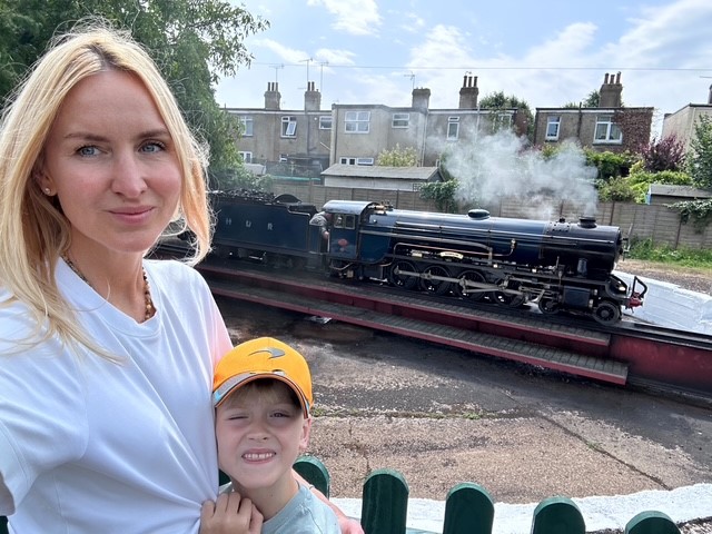 A mum stands on a train platform with her arm around a child in a yellow hat and a green steam train in the background. Landscape orientation