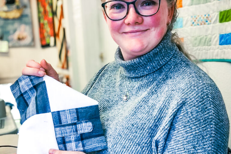 A girl in glasses (an Echoes sibling) is smiling at the camera. She's holding a blue and white patchwork square she has sewn for a memory blanket.
