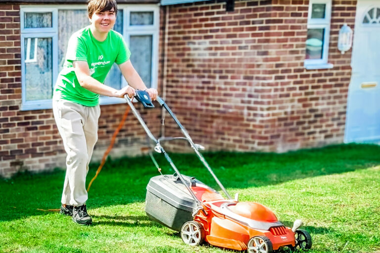 Momentum Young Ambassador in a green t-shirt mowing a lawn, smiling at camera.