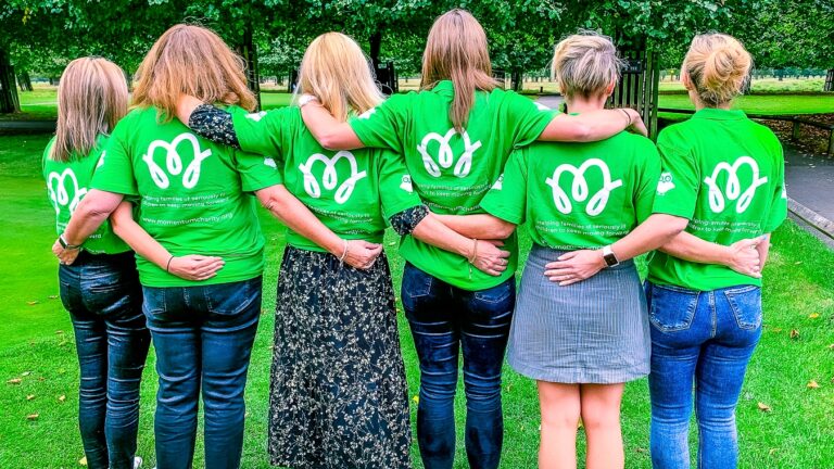 A group of Momentum staff, seen from behind with arms around each other's shoulders, wearing green t-shirts with the Momentum logo on the back, standing together in a park.