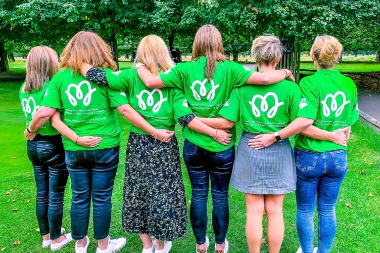 A group of Momentum staff, seen from behind with arms around each other's shoulders, wearing green t-shirts with the Momentum logo on the back, standing together in a park.