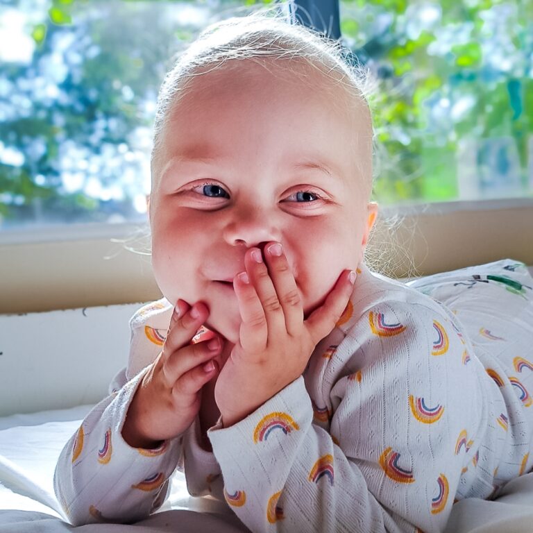 A supported child smiling at the camera with a hand covering their mouth playfully.