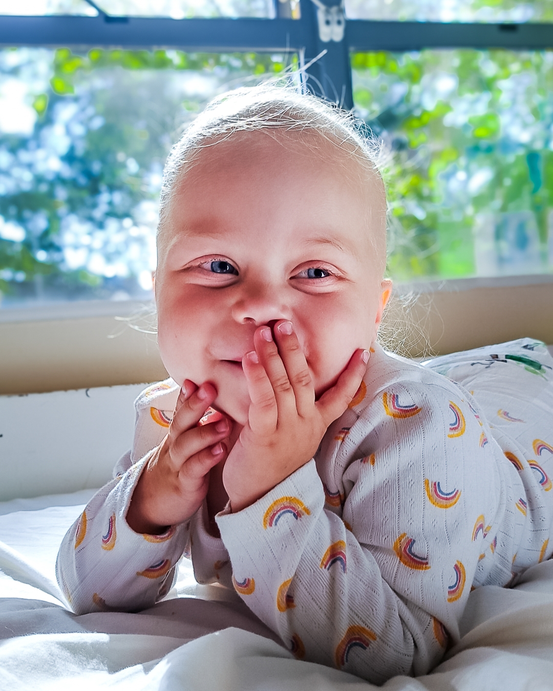 A supported child smiling at the camera with a hand covering their mouth playfully.