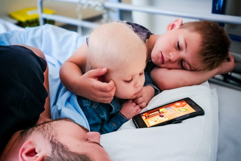 A supported child in hospital watching something on a parent's phone, with dad and brother lying beside her in the bed.