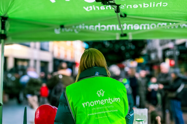 A mother supported through Momentum's Echoes programme fundraising in Kingston town centre for a Valentine's Day campaign, wearing a green tabard.