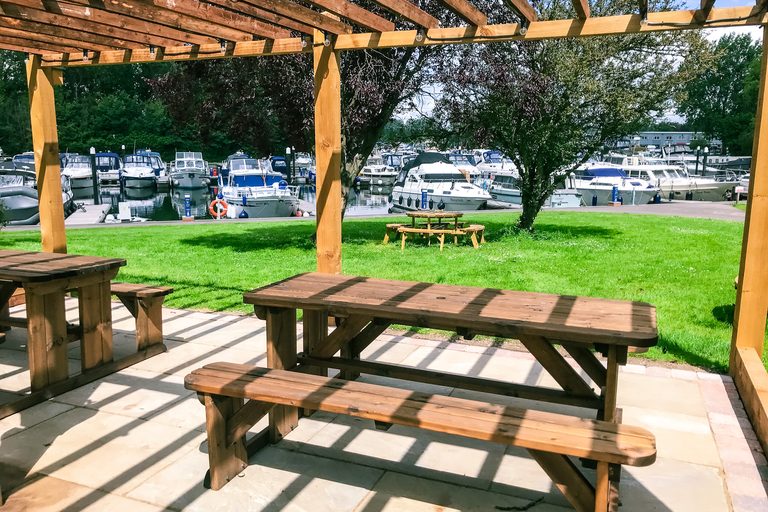 Shaded wooden pergola and picnic tables at the Shepperton Marina BBQ area overlooking moored boats.