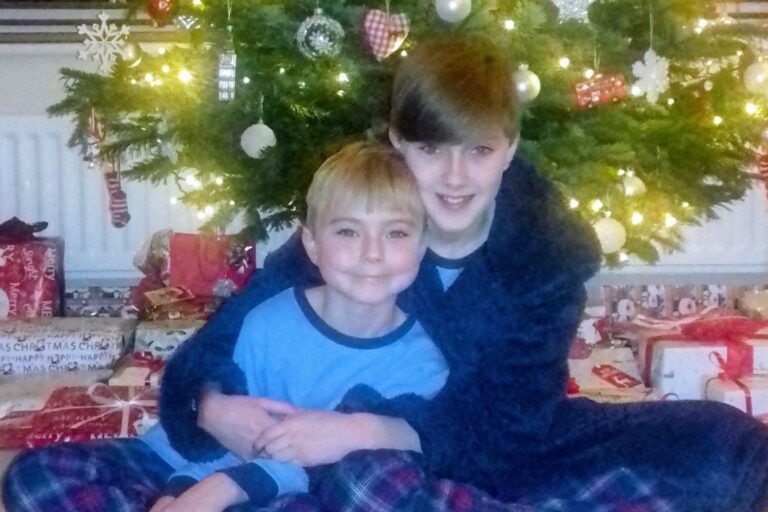 Two boys in PJs sit in front of a Christmas tree, hugging.
