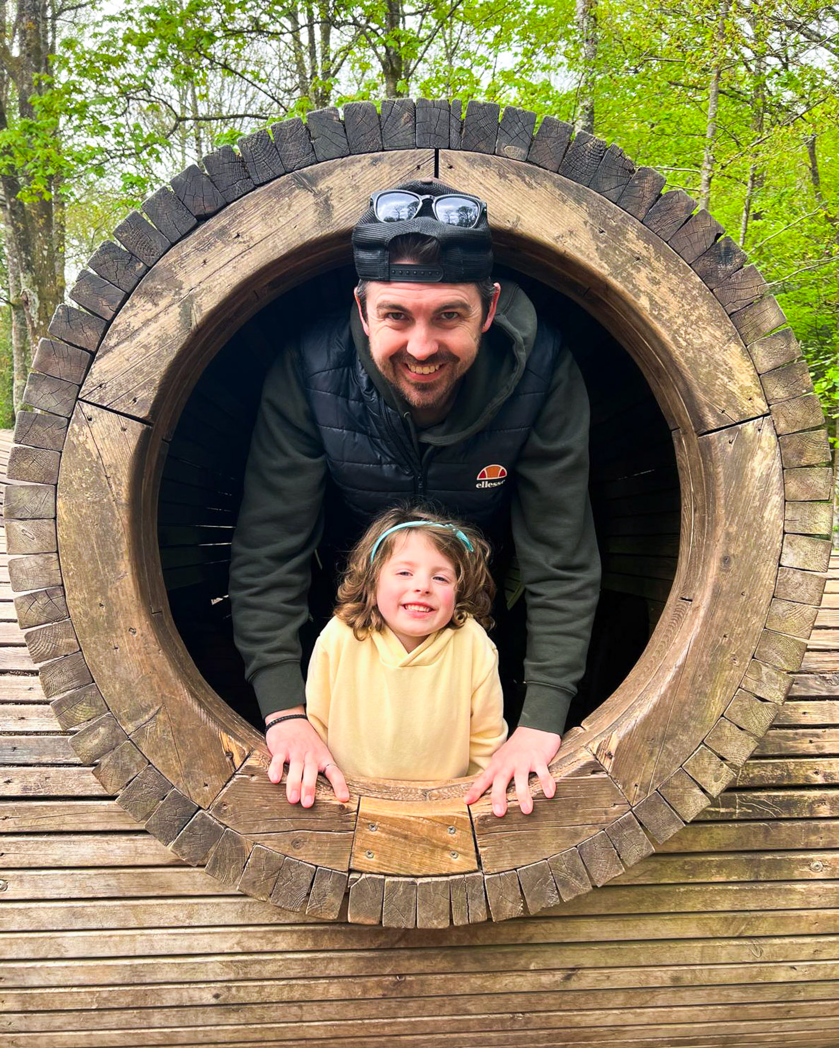 A Momentum-supported father and daughter look out through a round wooden tunnel.