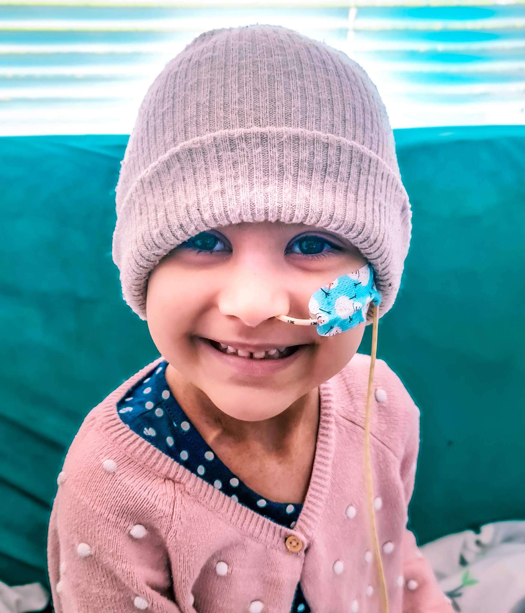 A child in a pale pink woolly hat and cardigan smiles at the camera - she has a nasal canula held in place by a blue plaster with snowmen.