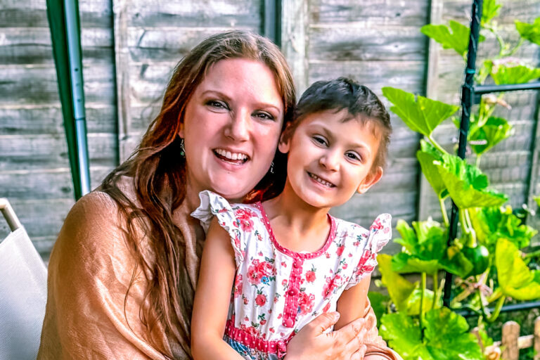 A mum sits on a garden chair with her child in a summer dress on her lap - both smiling at the camera.