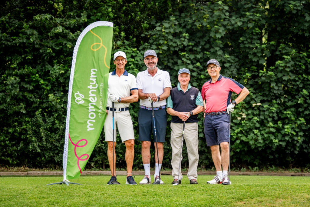 Momentum golf day partners dressed in gold attire holding their golf clubs pose beside the momentum branded flag on the golf course at Richmond Club
