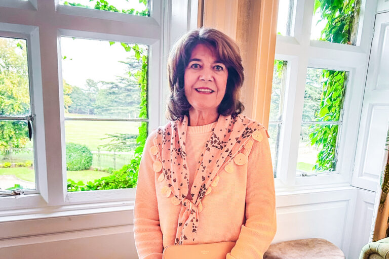 A woman stands in front of a brightly lit window wearing apricot and smiling