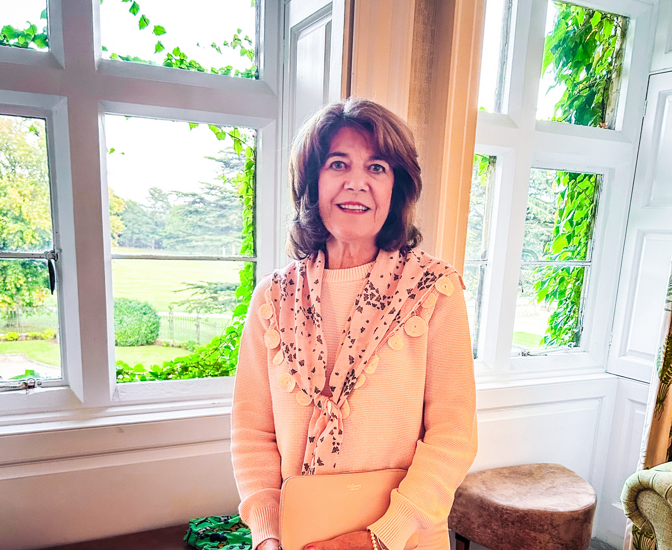 A woman stands in front of a brightly lit window wearing apricot and smiling