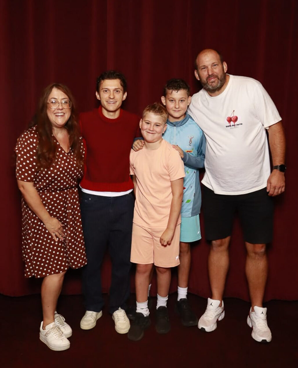 A family poses together for a photo with Tom Holland at a Spider Man film screening at Battersea Power Station. There is a red curtain backdrop.