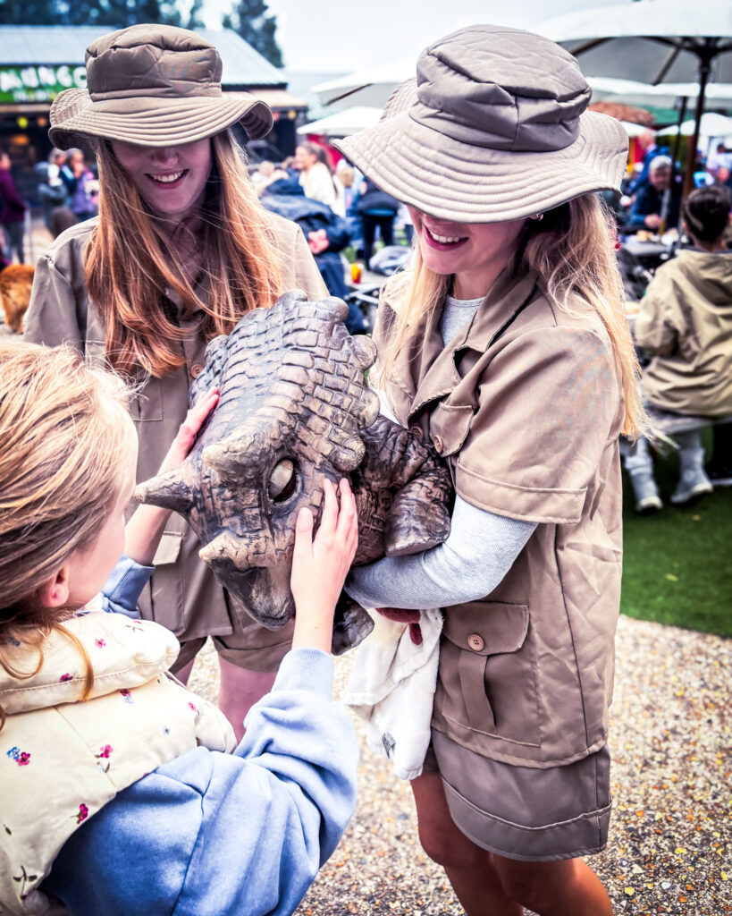 Two women dressed as safari explorers help children pet a lifelike dinosaur puppet at an outdoor family event.