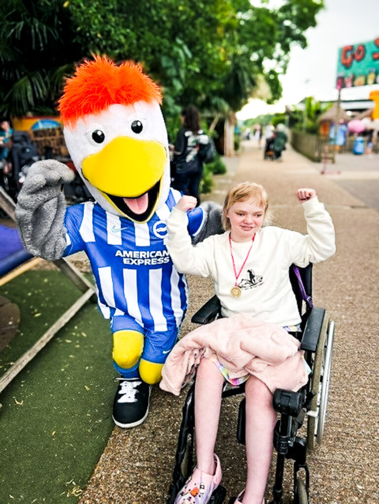 A girl wearing a medal sits in her wheelchair, fist bumping a football club's seagull mascot on a sunny match day out.