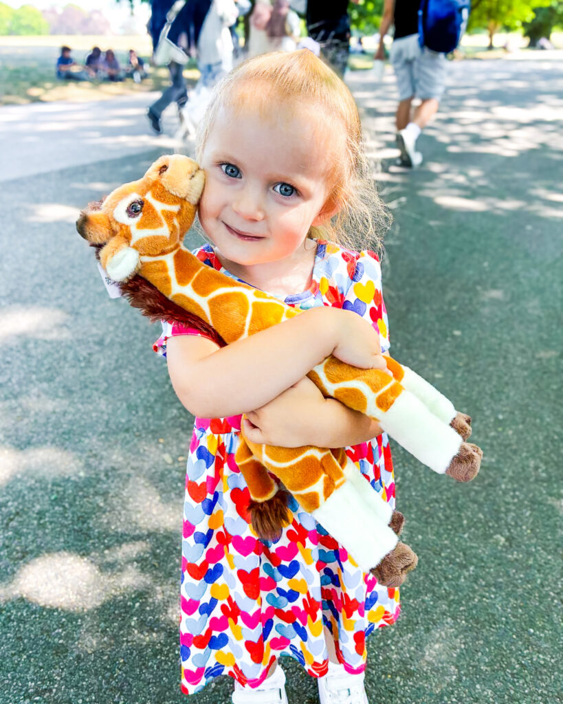 A toddler girl in a heart print dress hugs a plush giraffe toy tightly while walking through a sunny par