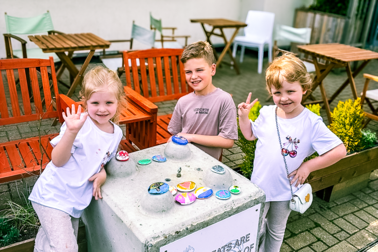 Three children - two girls and a boy smile as they paint pebbles at a Momentum event.