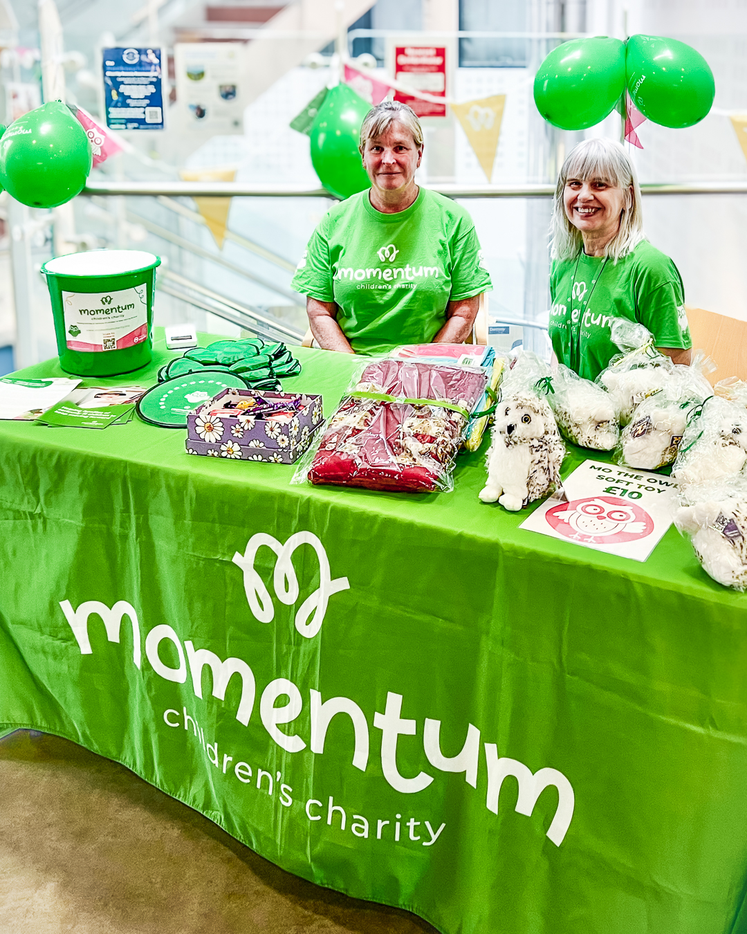 Two Momentum supporters smile at an event with balloons with a table full of soft toys and a collection bucket.