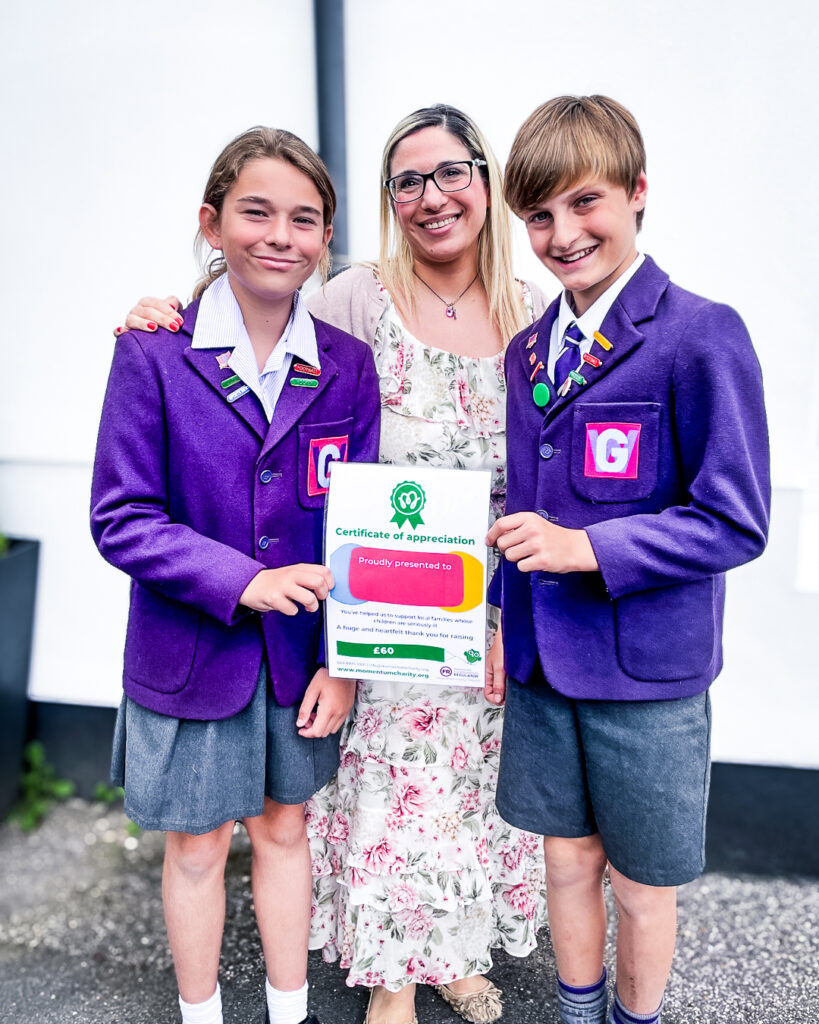 Two school aged children dressed in purple uniforms, stand smiling with a certificate with their Teacher after fundraising for Momentum. The certificate reads "Certificate of Appreciation - £60" from Momentum Children's Charity.