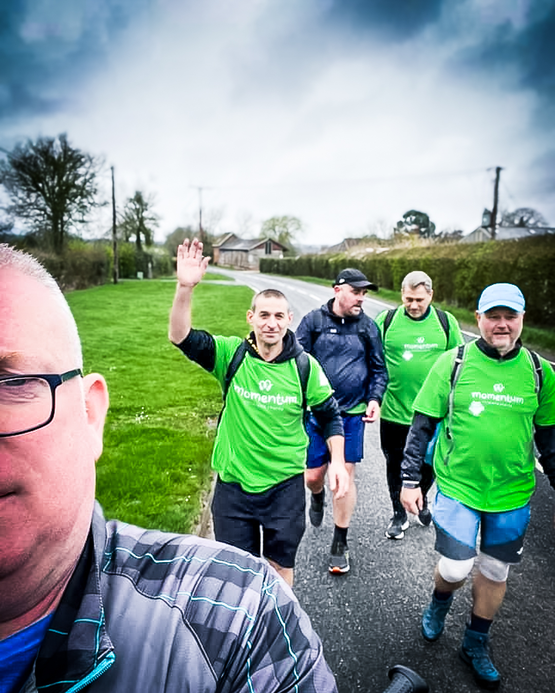 A sponsored walk selfie of 5 men. One man waves and the others are walking down a country lane while wearing Momentum green tshirts.