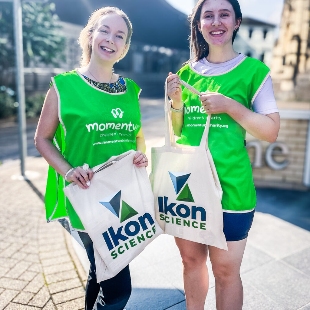 Two corporate partnership employees hold Ikon Science tote bags. They are smiling while looking directly at the camera in Momentum tabard tops.