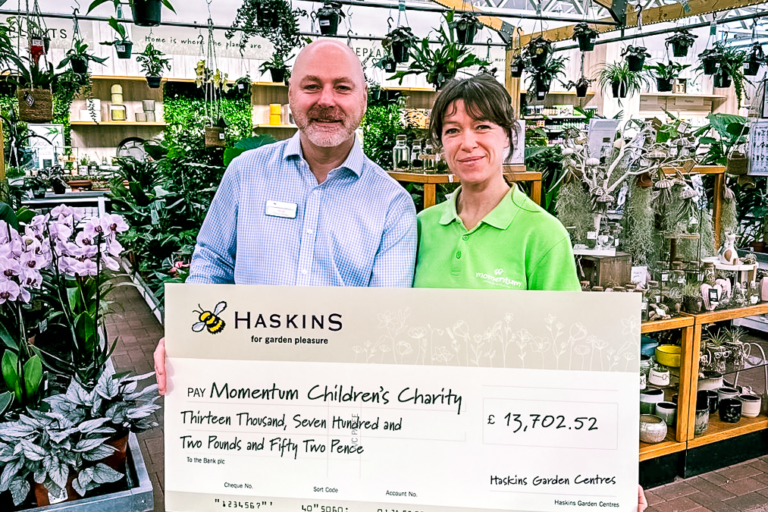 General Manager of Haskins Garden Centre stands holding a giant cheque alongside a Momentum Family Support Worker in amongst plants.