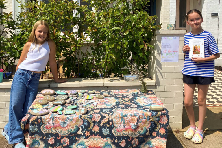 Two girls stand outside a table with pebbles they are selling to raise money for Momentum