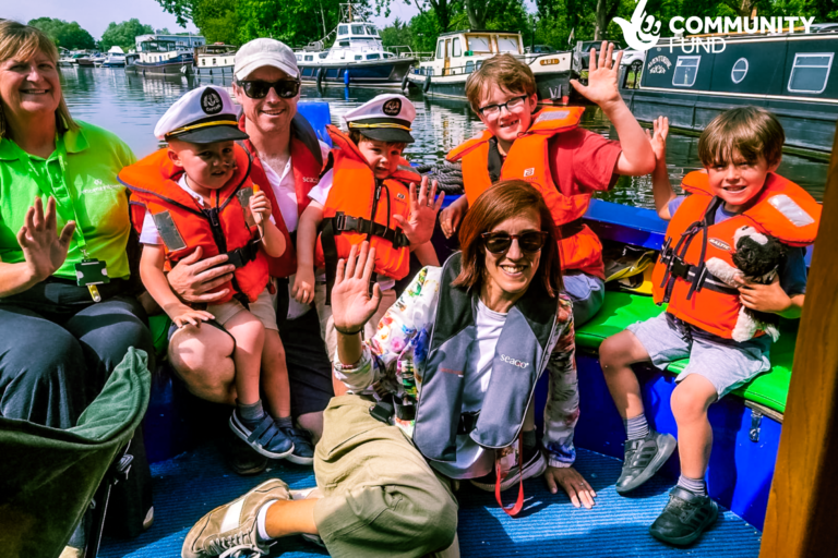 A supported family with four children in bright orange life jackets sit at the front of the Momentum boat waving at the camera. A Momentum family support worker sits beside them.