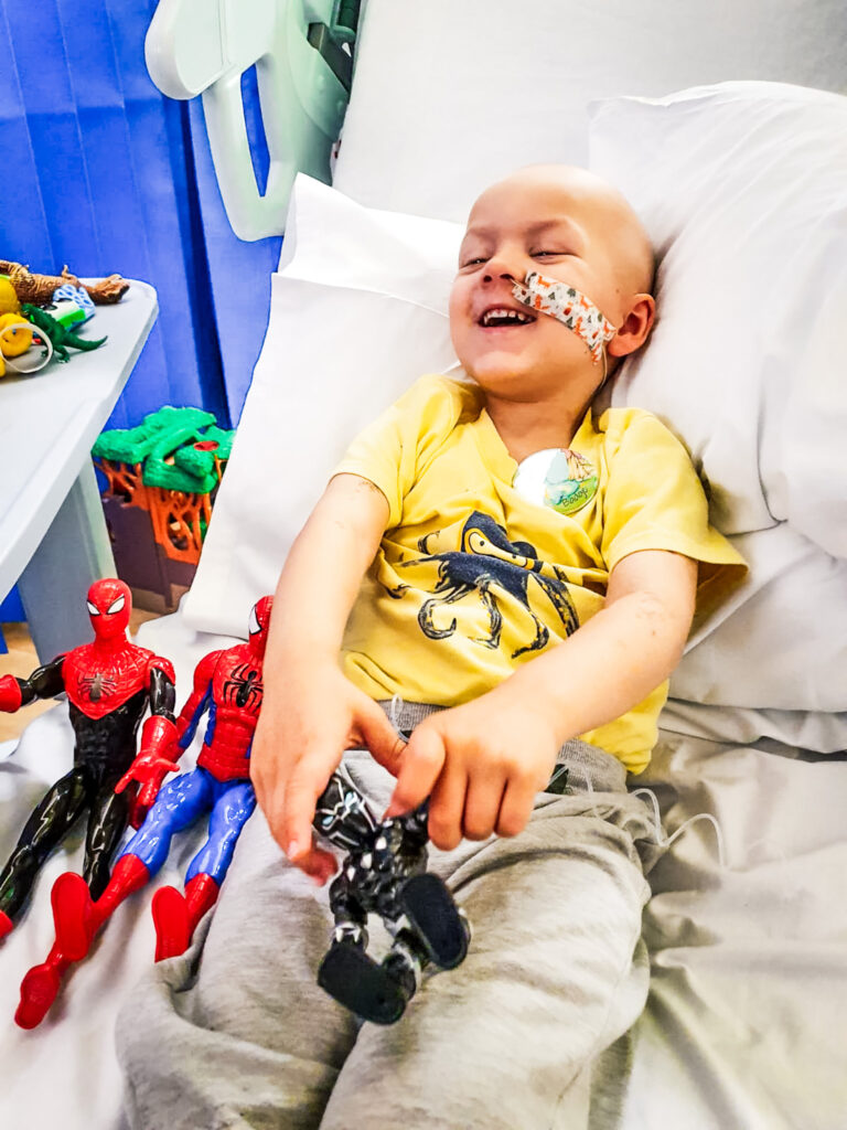 A small child smiles on a hospital bed while going through treatment with his toy spiderman dolls