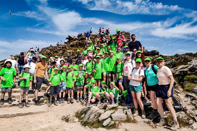 A large group of school children and their teachers in green Momentum t-shirts and hats stand on a rocky outcrop under a blue sky.