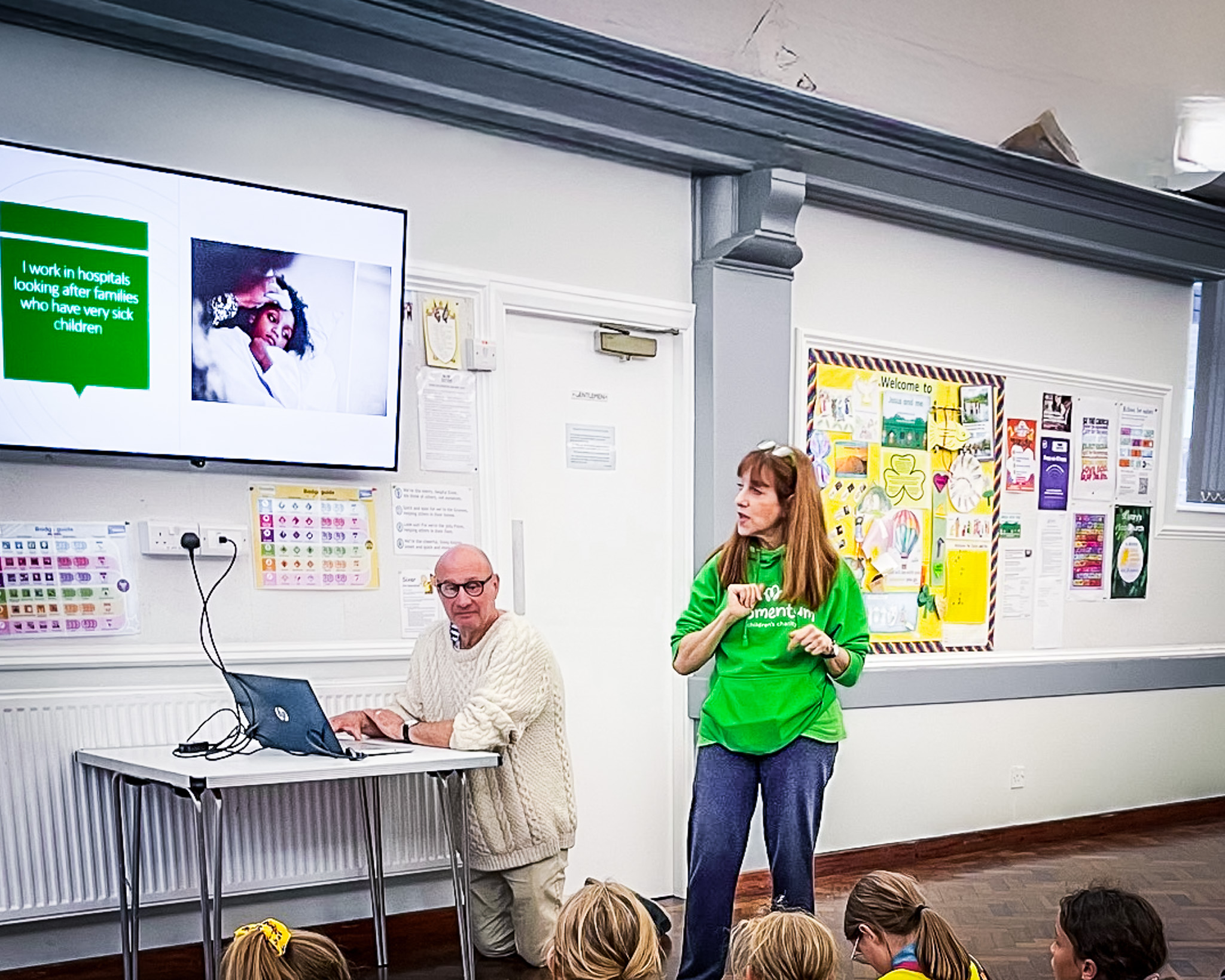 A Momentum worker in green sweatshirt looks at a screen showing slides as she gives a presentation about Momentum to a group of young Brownie Guides. A man kneels next to a laptop underneath the screen.