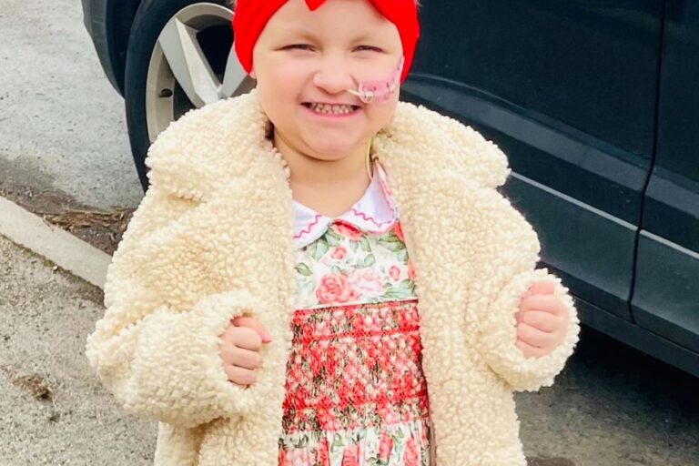 Little girl in a cream woolly coat, flowery dress and red bow in her hair. She has a nasal canula.