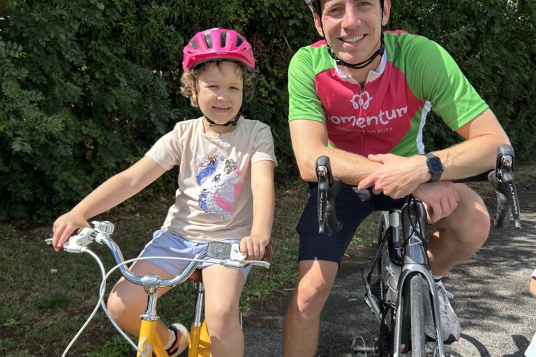 Man on a bike and Momentum cycle top smiles for camera together with a girl in a pink helmet on a yellow bike.