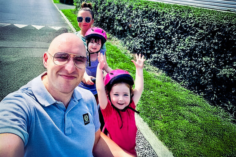 Dad in a blue top takes selfie along with his two young daughters in pink cycle helmets and mum in the back.