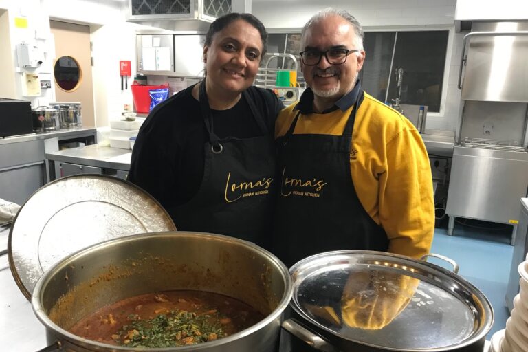 Two chefs in black aprons stand behind steaming pots of curry.