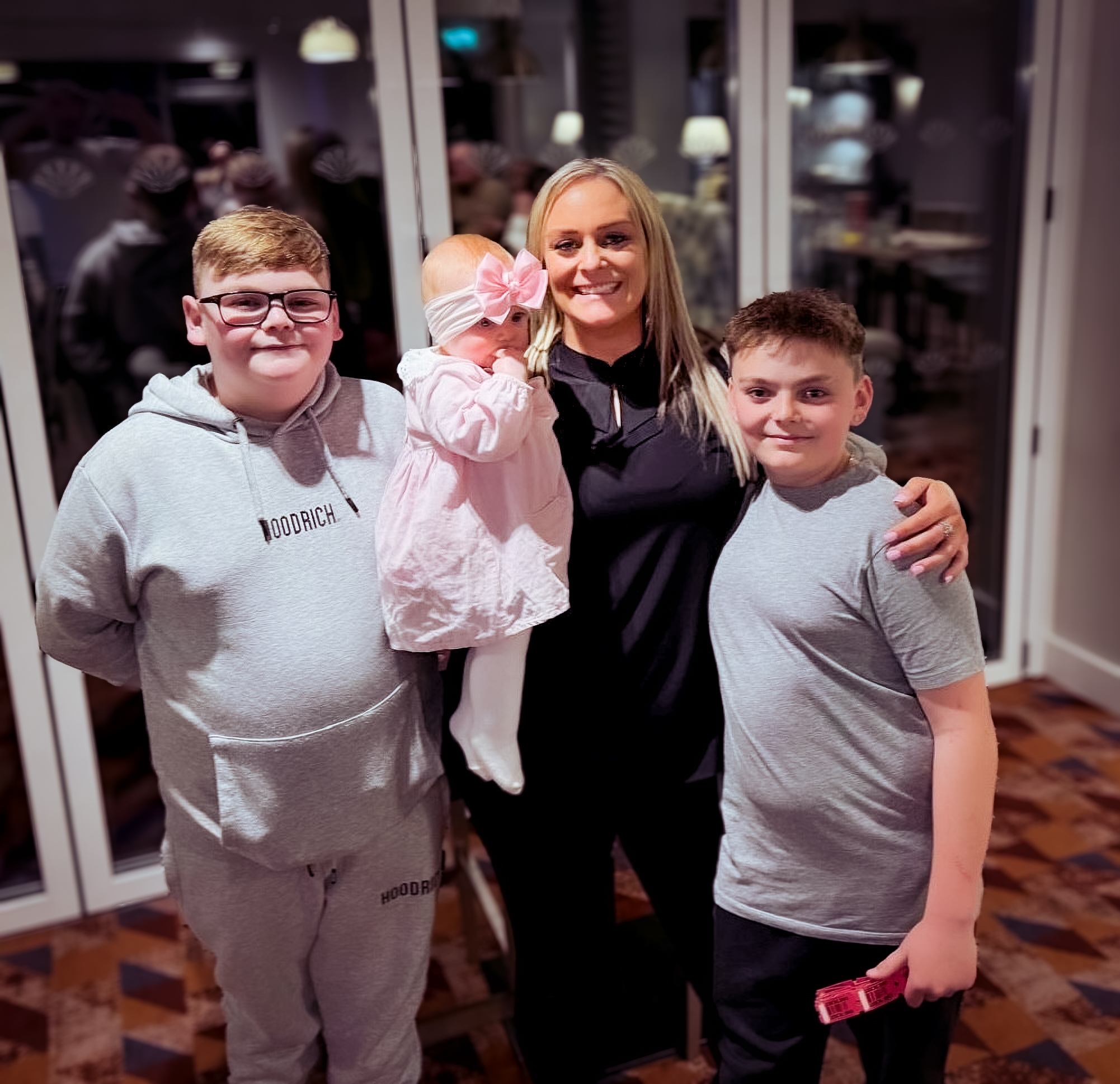 A Momentum-supported family stands in front of window at night time. Mum carries a toddler wearing a pink bow and dress and the boys are in grey.