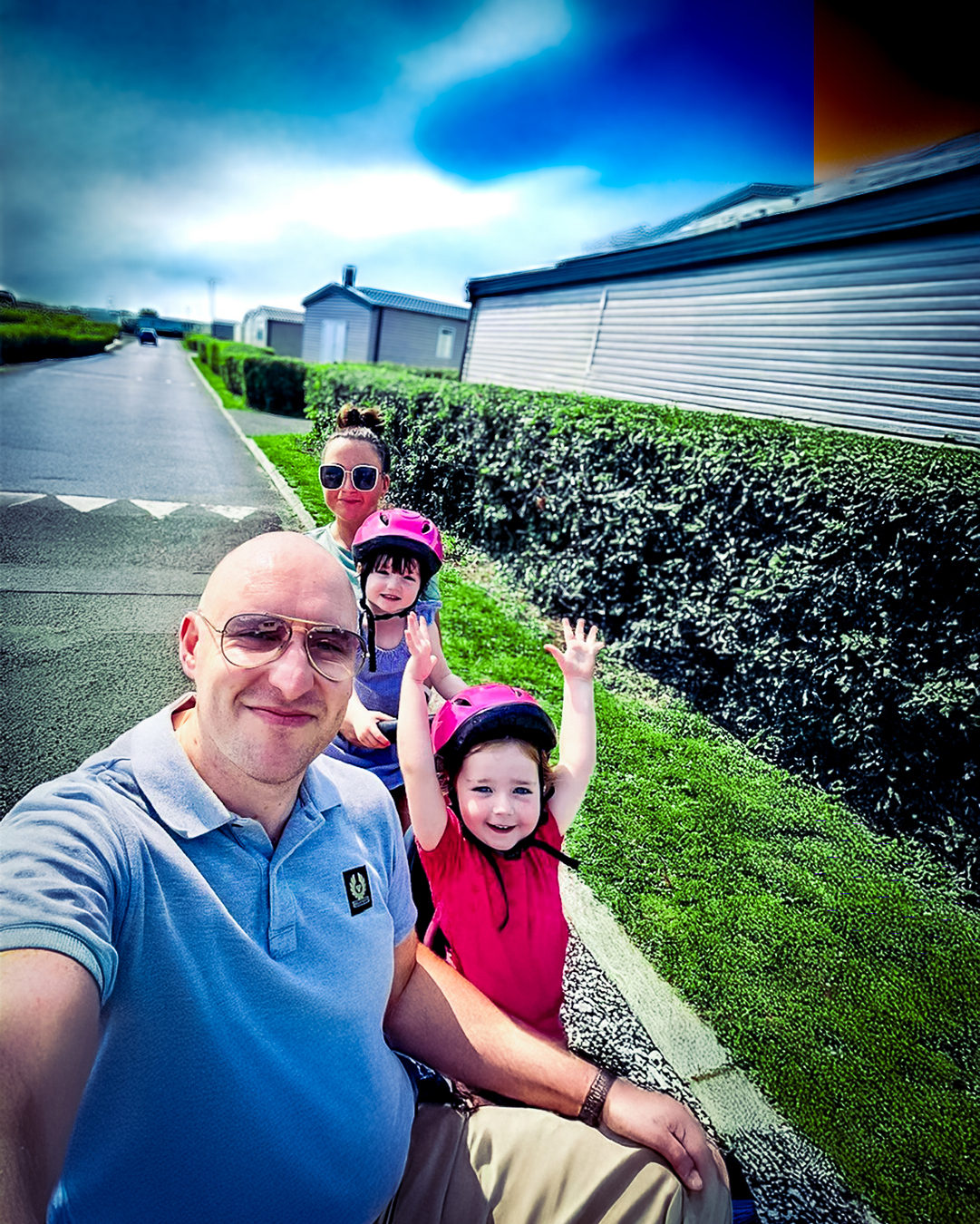 A family of four take a selfie outside in a holiday park, the two kids are in pink cycle helmets and one raises her hands in the air.