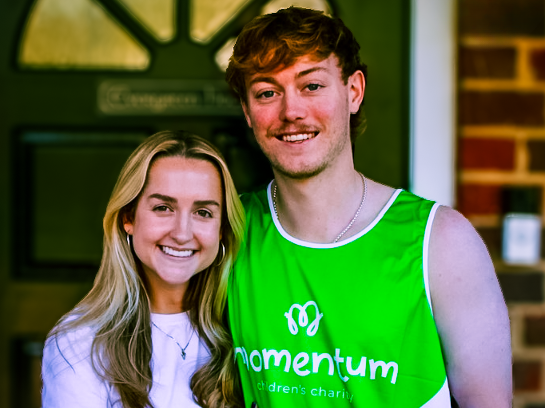 One fundraiser preparing to run the Florence marathon for Momentum Children's Charity. He is stood by his partner. They both smile at the camera. He is wearing his Momentum running vest.