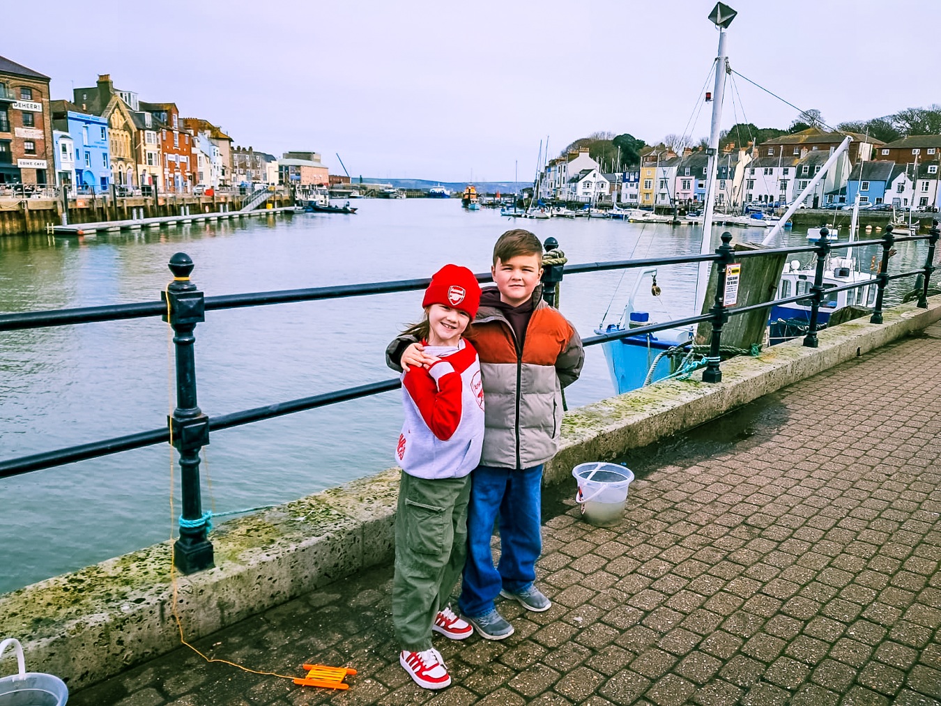 Two siblings stand together on a quay in a quaint fishing village with colourful cottages and fishing boats behind. They have crabbing lines and buckets at their feet.
