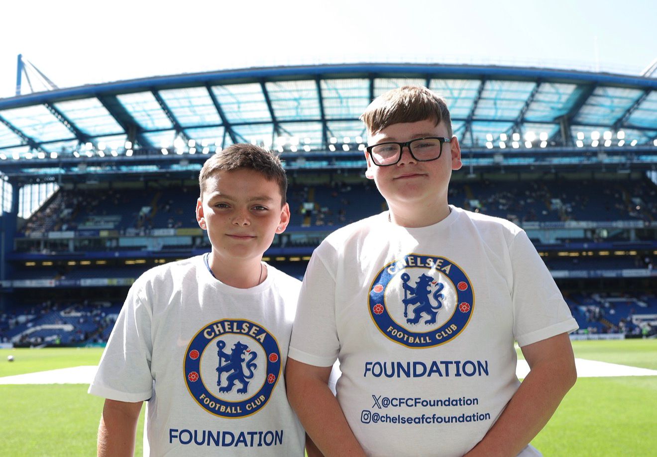 Two siblings in Chelsea Foundation t-shirts stand together on the pitch in the Chelsea stadium.