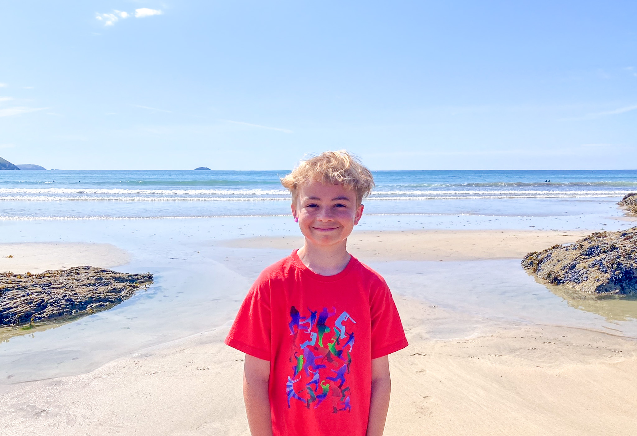 A boy in a red t-shirt stands on a sandy beach with the sea in the background, smiling broadly at the camera.